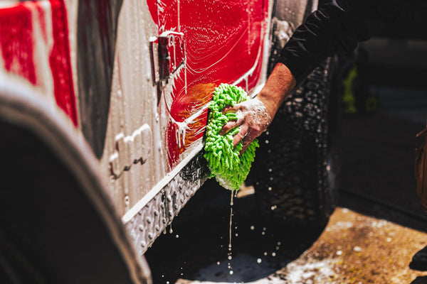 Person washing a red vehicle with a green microfiber wash mitt, focusing on the lower side panel, with the vehicle covered in soap suds.