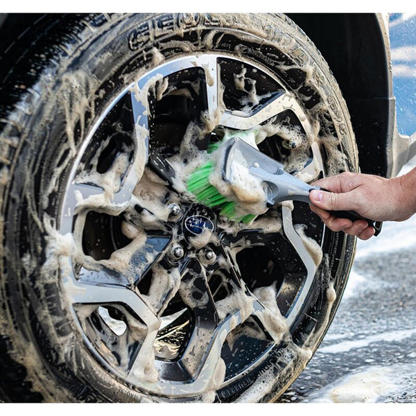A hand holding a short green Wheel and Body brush cleaning a foamy car tire rim.