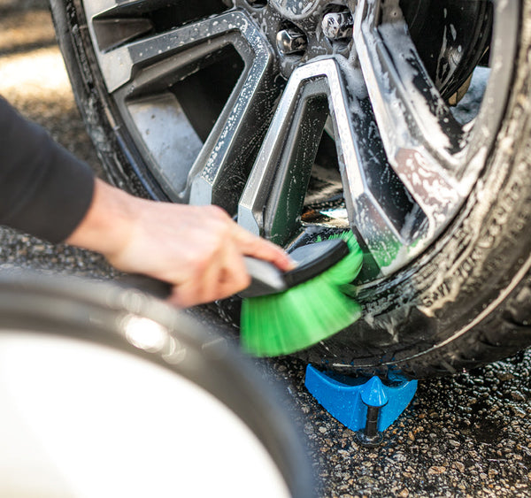 A hand holding a short green Wheel and Body brush cleaning a foamy car tire rim.
