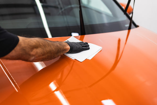 A closeup of a person cleaning the hood of an orange car with a while paper towel.