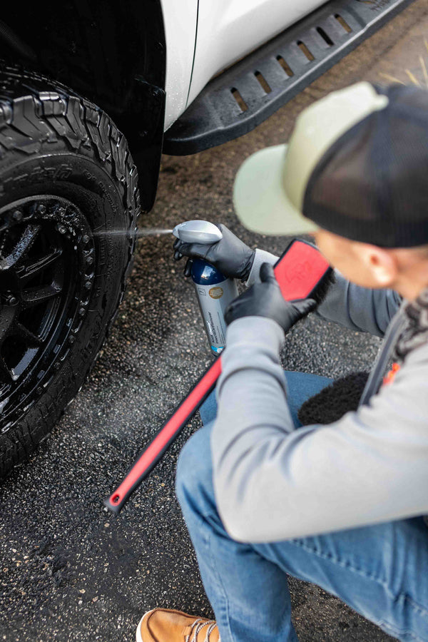 A person holding a Detail Factory Fender Well Brush Red and spraying a KochChemie product on the tire of a white truck.
