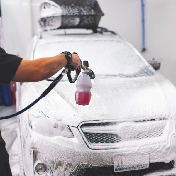 Person using a foam cannon to apply soap onto a car during a wash, with the car fully covered in foam.