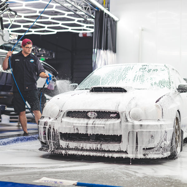Person using a foam cannon to apply soap onto a car during a wash, with the car fully covered in foam.