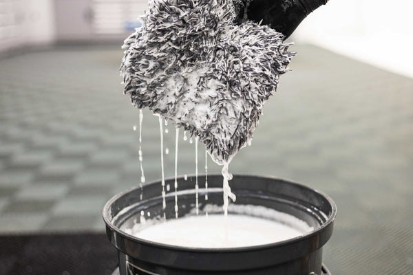 A black and white microfiber wash mitt, dripping soapy water into a black bucket filled with water and Koch-Chemie Ceramic Effect shampoo.