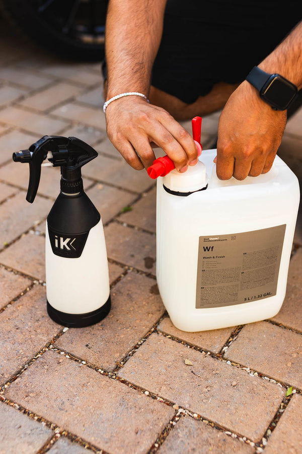 A person is attaching a red dispenser to a 5-liter container of Koch Chemie Wash & Finish, with a white and black iK spray bottle placed nearby on a paved surface.