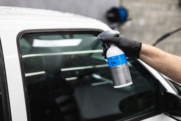 A gloved hand spraying KochChemie Clay Spray onto the window of a white vehicle.