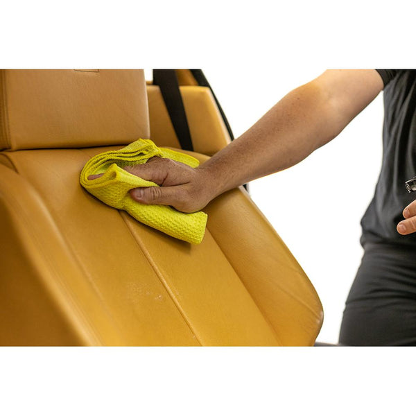 A person wiping a mustard colored leather seat with a yellow standard waffle weave towel.