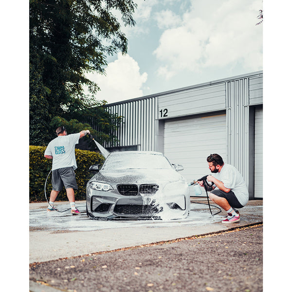The same person detailing a car outdoors with a foam cannon and a hose.