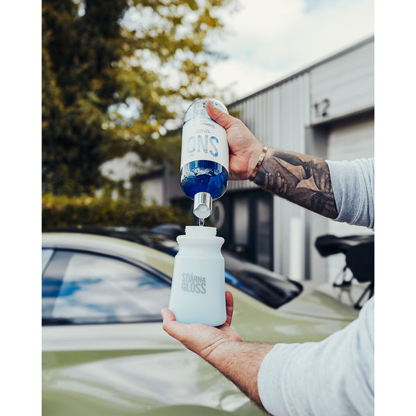 A closeup of a person pouring Stjarnagloss Sno into a foam cannon with a green car in the background.