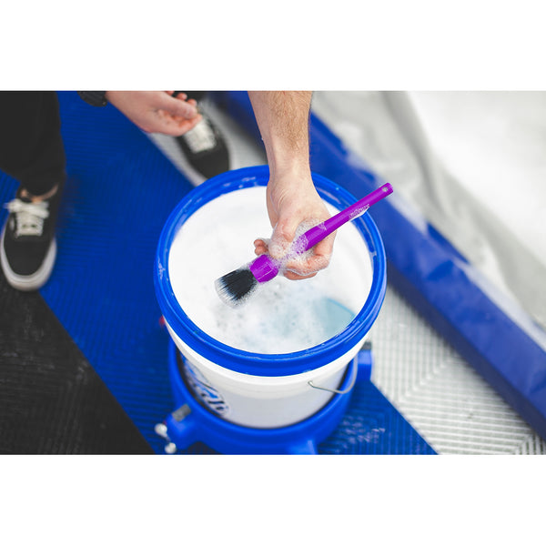 A person holding a purple detailing brush dipped in a white bucket filled with soapy water, preparing to clean a vehicle.