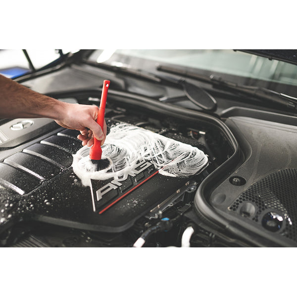 A closeup of a person cleaning an engine cover with a red synthetic detailing brush.