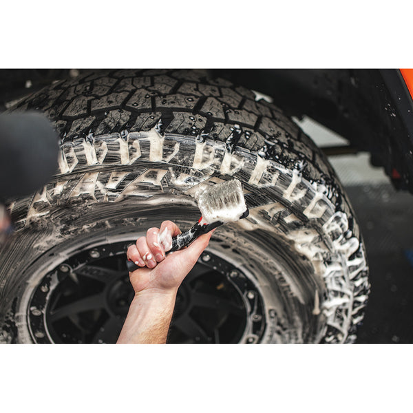 A person showing a red Detail Factory Tire Scrub Brush in front of a tire covered in foam.