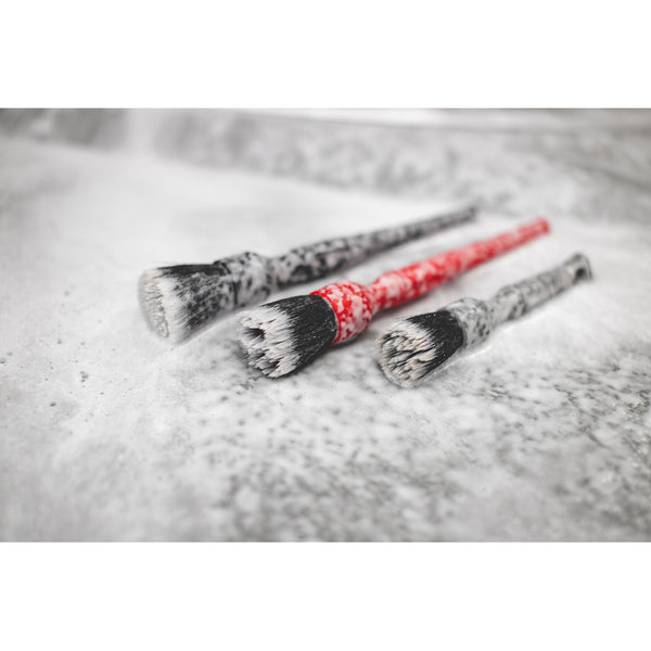 A black, red, and grey synthetic detailing brush in parallel covered in foam.