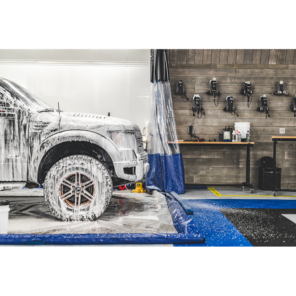 A white truck covered in foam inside a car detailing bay with various cleaning tools and equipment mounted on the wall in the background.