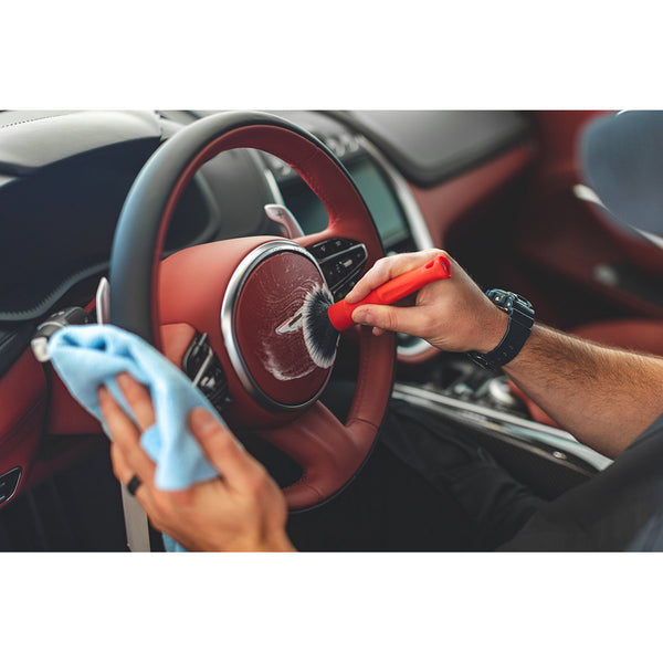 A person using the Detail Factory red Synthetic Detailing Brush to clean a maroon leather steering wheel.