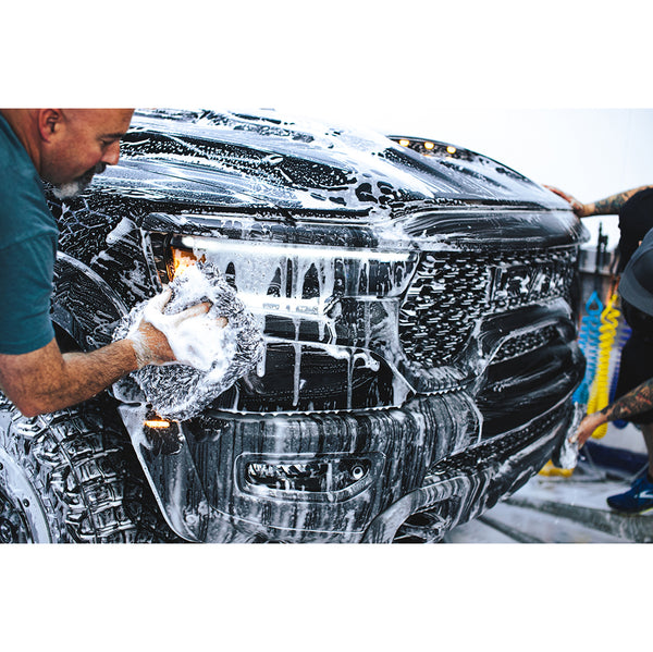 The front of a black truck covered in foam while to people scrub it with long fiber towels.