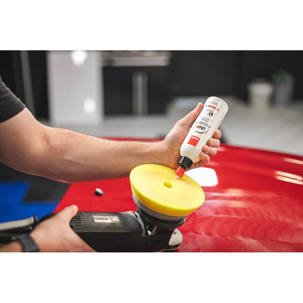 A closeup of a person pouring Rupes Uno Protect onto a polisher with a red car in the background.