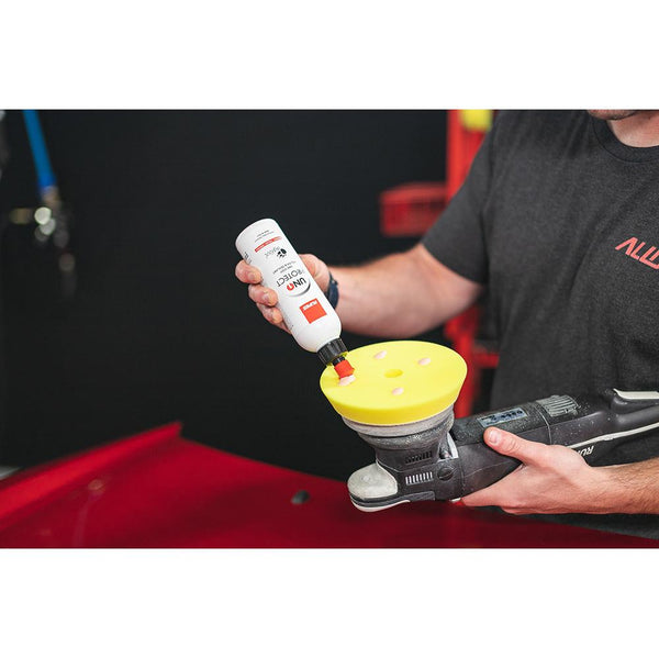 A closeup of a person pouring Rupes Uno Protect onto a polisher with a red car in the background.