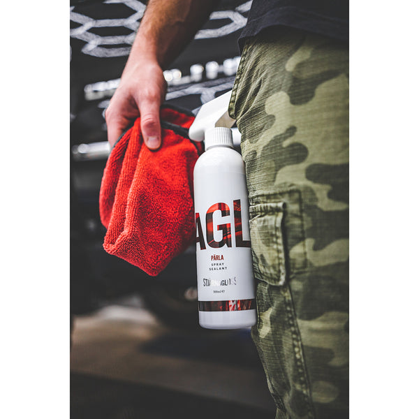 Close-up of a person holding a red microfiber towel and a bottle of Stjärnagloss Pärla spray sealant, with camo-patterned pants visible in the foreground.