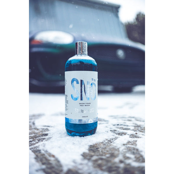 A bottle of Stjarnagloss Snow Foam on concrete covered in snow with a green car in the background.