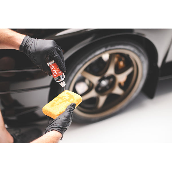 A closeup of a person wearing black rubber gloves pouring Gtechniq Exo onto a yellow pearl sponge with a background of a sports car.