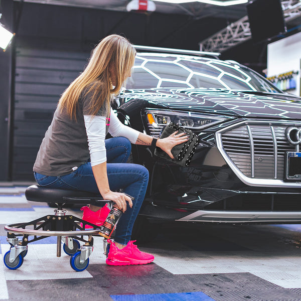 A woman is sitting on a rolling stool, detailing the front headlight of a black car using a Wolfpack towel by The Rag Company. She is holding a spray bottle in her right hand and scrubbing the headlight with the towel in her left hand. The garage setting features a clean, organized workspace with a hexagon light pattern reflecting on the car's hood. She is wearing a gray shirt, jeans, and bright pink sneakers.