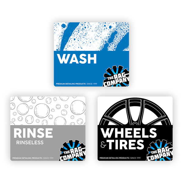 Three product labels from The Rag Company for car detailing categories. The first label, in blue, reads 'Wash' with a design of water splashes. The second label, in gray, reads 'Rinse Rinseless' with a bubble pattern. The third label, in black and white, reads 'Wheels & Tires' with an illustration of a wheel. All labels include The Rag Company logo and the tagline 'Premium Detailing Products | Since 1999' at the bottom.