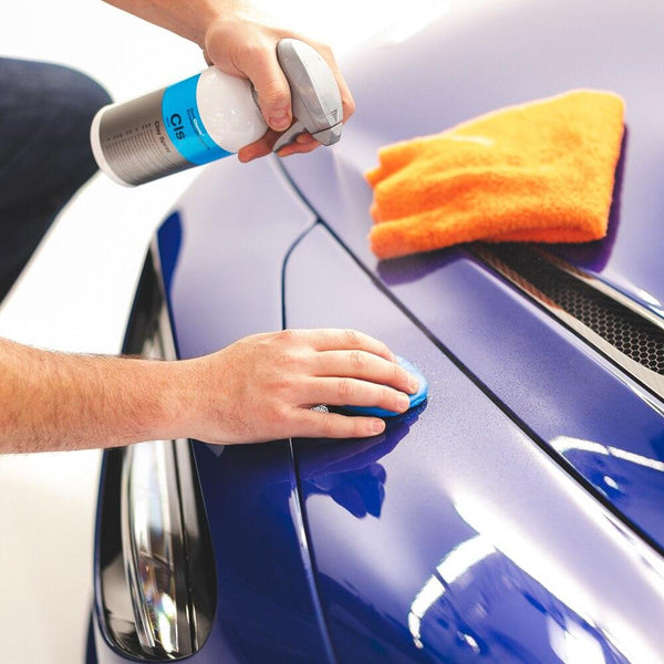 A closeup of a person spraying Koch Chemie Clay Spray onto a Clay Bar on a blue car.