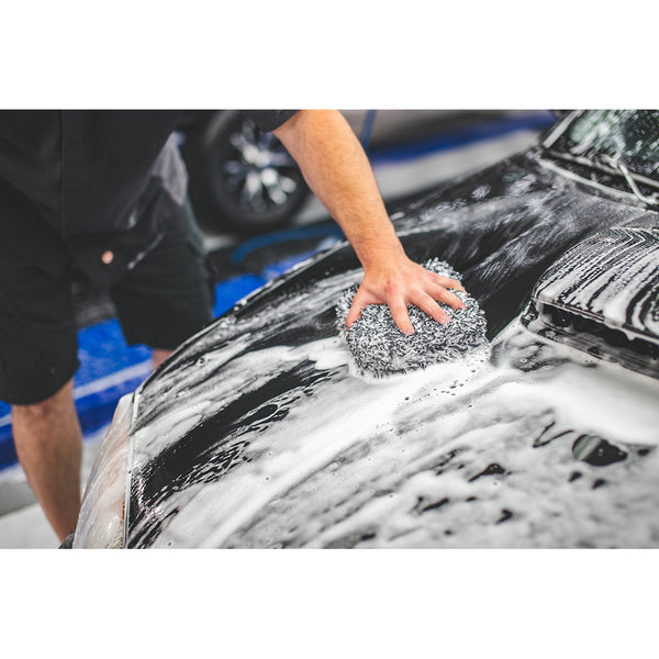 A closeup of someone using The Rag Company Ultra Wash Mitt on the hood of a black car covered in foam.