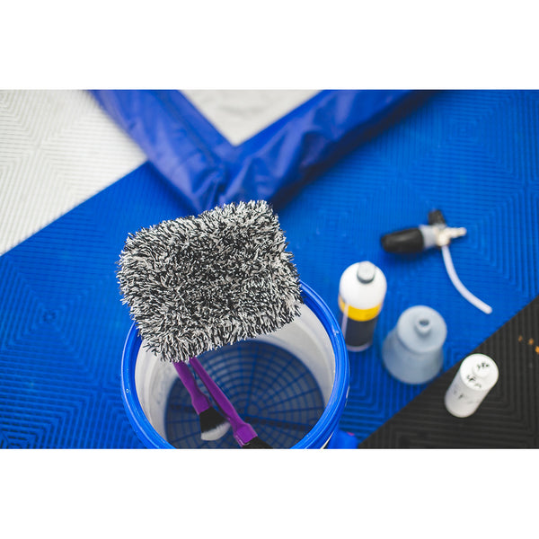 A close-up view of a bucket filled with water and a microfiber wash mitt resting on top. The bucket is placed on a blue mat with a few car cleaning bottles and a spray nozzle around it, ready for use in a detailing process.