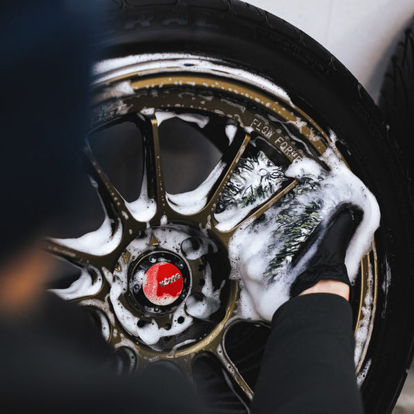 A closeup of someone wearing a black rubber glove cleaning a foam covered car tire rim with an Ultra Wheel Mitt.