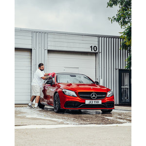 A person wearing all white clothes drying an orange Mercedes with a red towel.