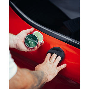 A person scrubbing a red car with a round scrubber in their right hand and holding Stjarnagloss Fantastisk in the left hand.