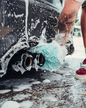 A person using a teal towel drenched in foam to clean the exhaust pipe of a black car covered in foam. 