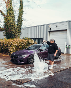 A person wearing a black hoodie and gray shorts using a pressure washer to clean a purple car, with soap suds splashing from a bucket labeled 'STJARNAGLOSS' in an outdoor setting near a building with a garage door.