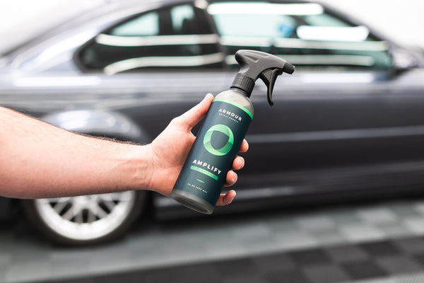 A person holding a spray bottle of Armour Amplify in front of a grey car.