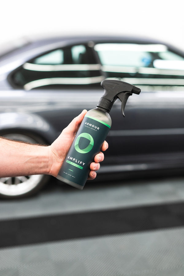 A person holding a spray bottle of Armour Amplify in front of a grey car.