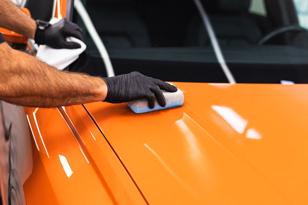 A closeup of a person cleaning the hood of an orange car with a blue and grey applicator sponge.