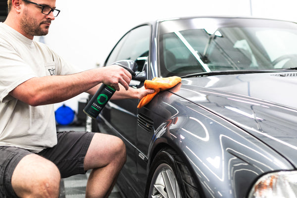 A person spraying Armour Ceramic Spray Sealant onto a grey car and wiping it with an orange towel.