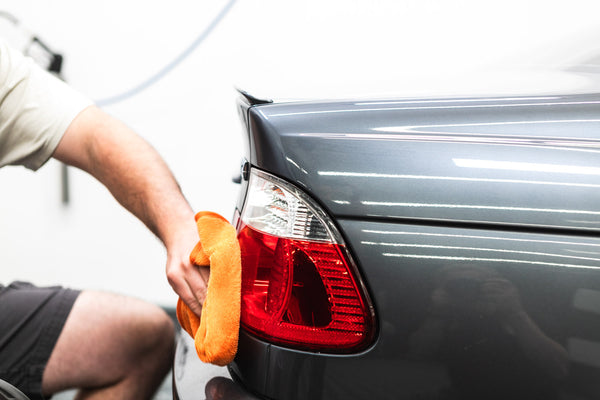 A closeup of a person wiping the back headlight of a grey car with an orange towel.