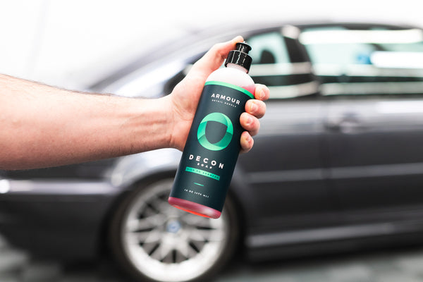 A person holding a bottle of Armour Decon Soap in front of a grey car.