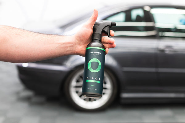 A closeup of a person holding a spray bottle of Armour Pilot in front of a grey car.