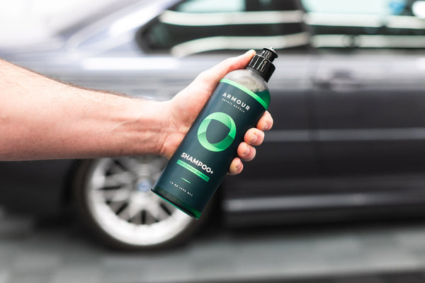 A person holding a bottle of Armour Shampoo+ in front of a grey car.