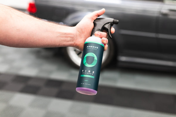 A person holding a spray bottle of Armour Tire Cleaner in front of a grey car.