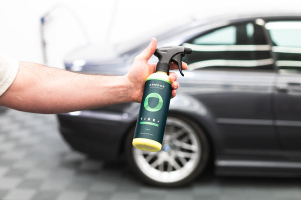 A closeup of a person holding a spray bottle of Armour Tire+ in front of a grey car.