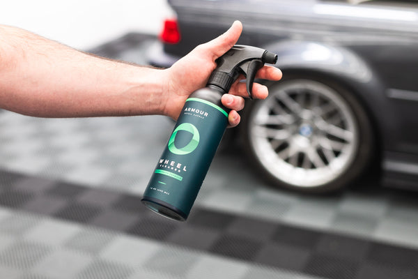 A closeup of a person holding a spray bottle of Armour Wheel Cleaner in front of a grey car.