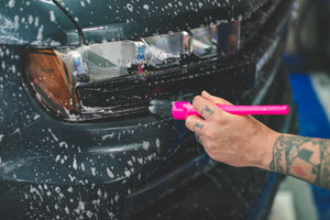 A person using a pink synthetic brush to clean the front of a black truck.