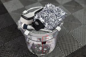 Detail Factory Clean Screen Grey placed on top of a clear wash bucket, holding detailing brushes and a microfiber wash mitt.