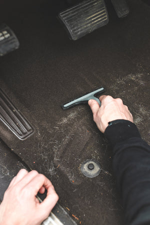A gloved hand using a Detail Factory Pet Hair Rake on the carpeted ground of a vehicle.