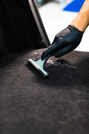 A gloved hand using a Detail Factory Pet Hair Rake on the carpeted ground of a vehicle.